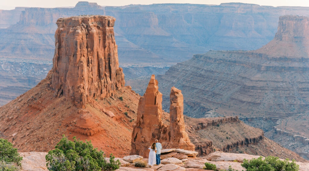 Bride & groom stand on the edge of a cliff overlooking the remote desert of Moab, Utah.