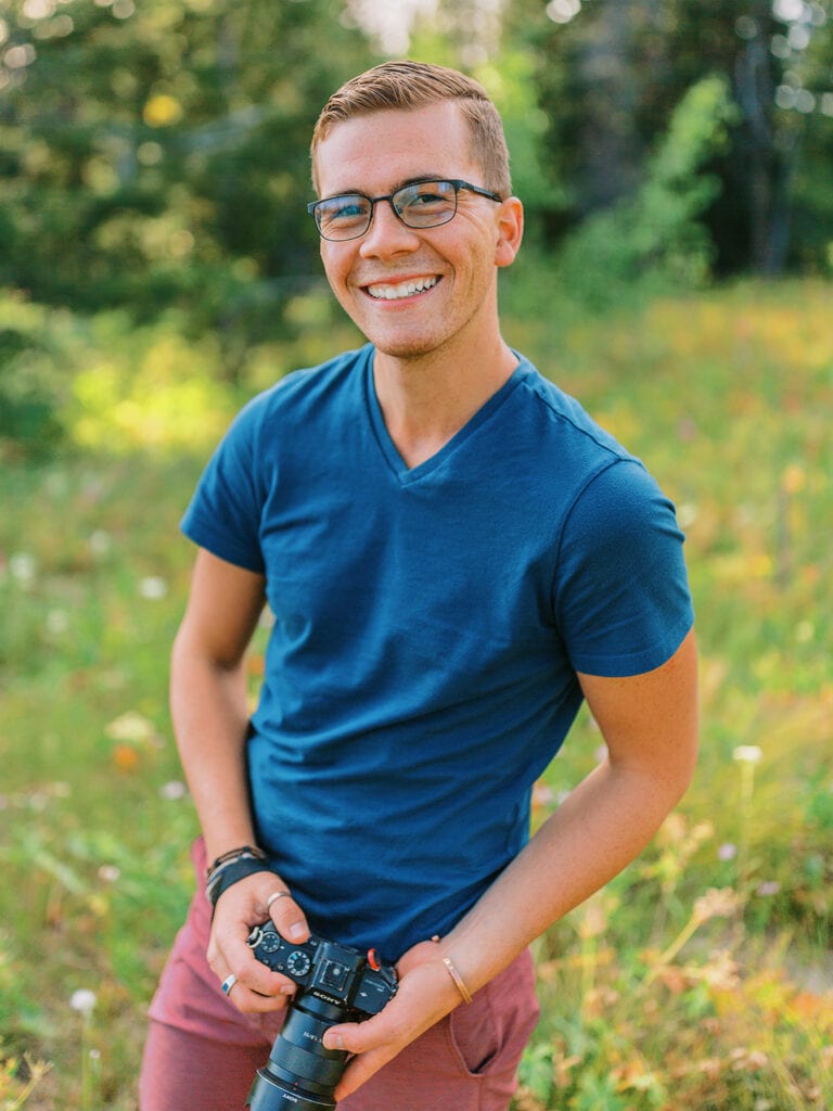 Malachi Lewis, a photographer, smiling and holding a camera in mountain settings with wildflowers in the background.