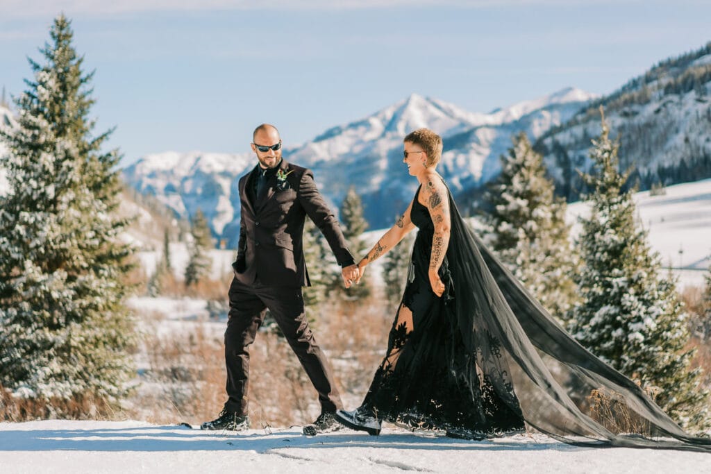 Bride in a black dress hikes with her groom during their snowy elopement in Colorado.