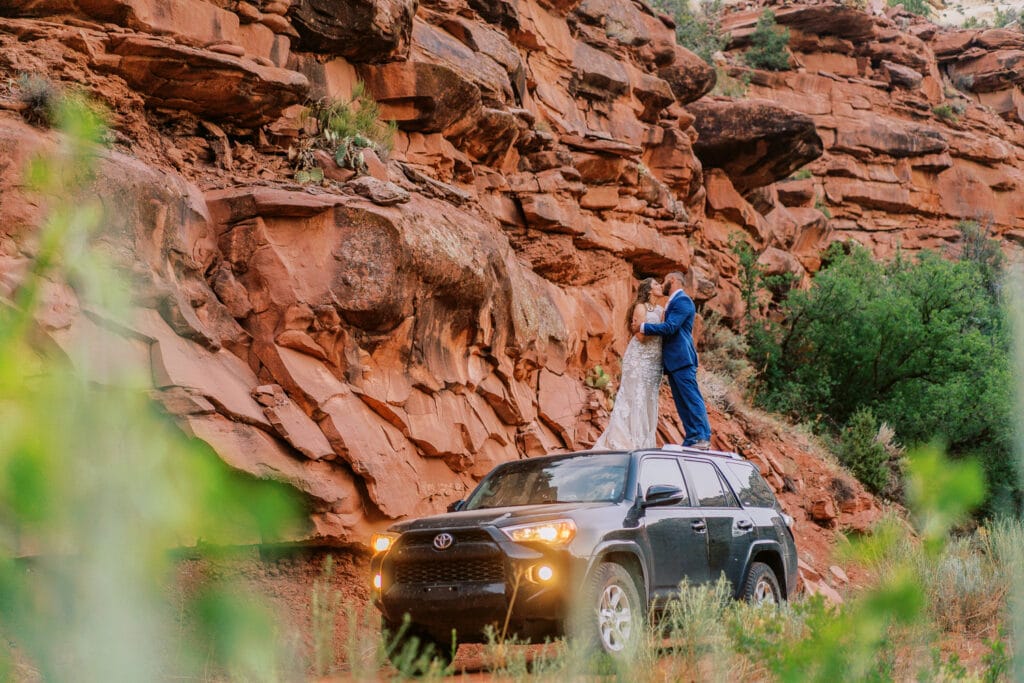While surrounded by red rock in a canyon, a couple kisses while standing on top of their 4Runner.