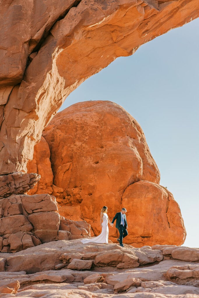 Couple walks under North Window Arch during their wedding day in Arches National Park.