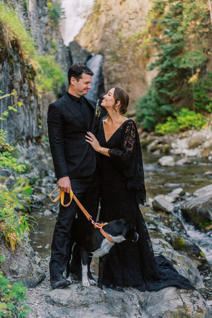 Bride in a black dress smiles at the groom during their waterfall elopement in Colorado.