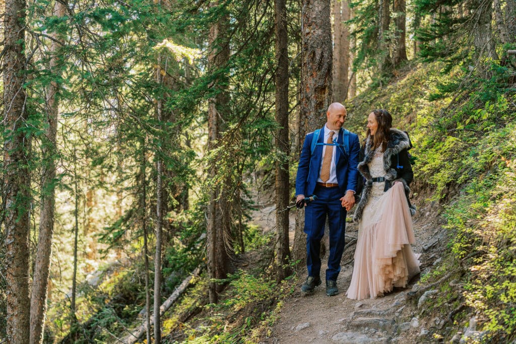 Bride in a pink dress and groom in a blue suit hike down a trail in the forest during their wedding day in the mountains.