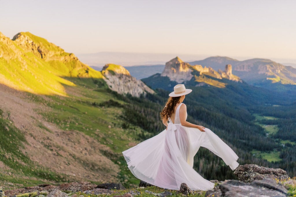 Bride in a hat twirls around a mountain overlook at sunrise.