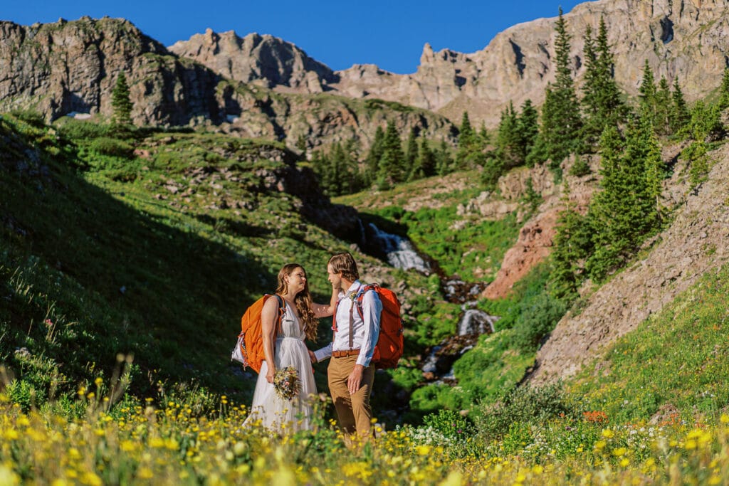 Bride and groom standing in front of a waterfall in the San Juan Mountains with yellow wildflowers in the foreground.
